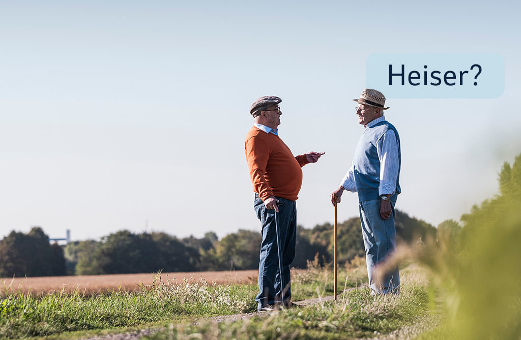 Zwei ältere Männer stehen sich auf einem Feldweg gegenüber und unterhalten sich. Der Mann rechts stützt sich auf einen Gehstock. Über ihm ist eine Sprechblase mit dem Wort „Heiser?. Im Hintergrund ist eine Landschaft mit Bäumen und Feldern zu sehen.