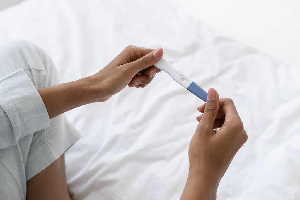 Eine Frau sitzt im Schneidersitz auf einem Bett und hält einen Schwangerschaftstest in ihren Händen