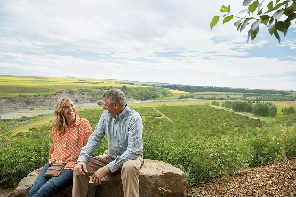 Ein Mann und eine Frau sitzen auf einem großen Stein vor einer grünen Landschaft. Sie sehen sich an und lächeln.