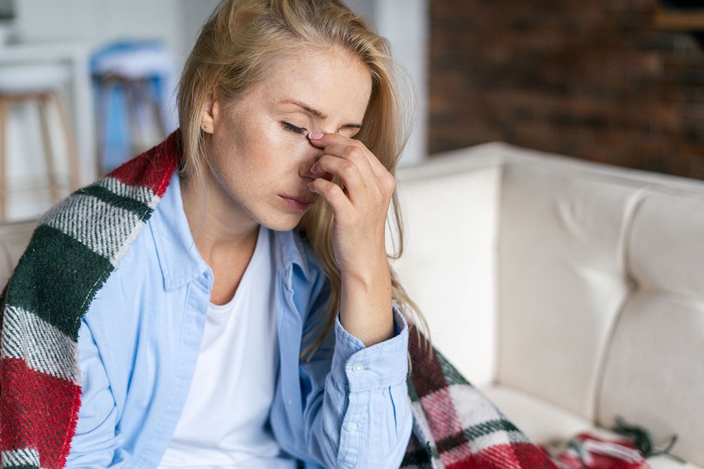 Eine blonde Frau sitzt auf einem Sofa mit Decke um die Schultern. Sie hat die Augen geschlossen und stützt ihren Kopf mit der Hand ab.