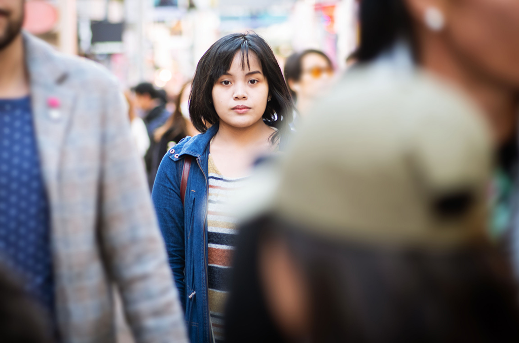 Eine junge Frau asiatischer Herkunft blickt in die Kamera, viele unscharf zu erkennende Menschen gehen an ihr vorbei.