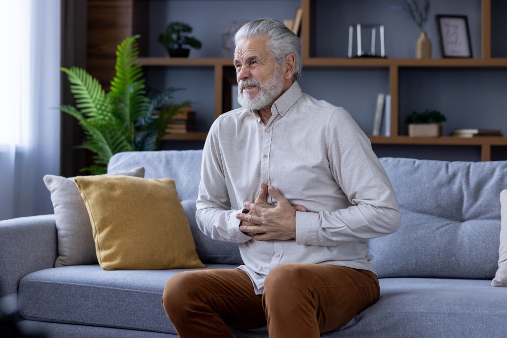 Die Abbildung zeigt einen älteren Mann mit grauen Haaren und Bart, der auf einem Sofa sitzt. Er hält sich mit beiden Händen den Bauch und hat einen schmerzverzerrten Gesichtsausdruck.