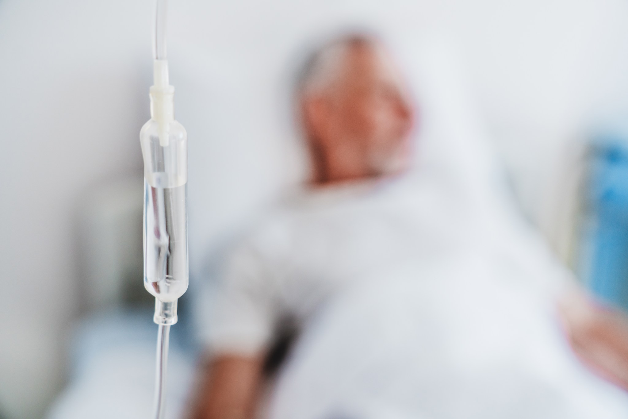Das Bild zeigt die Nahaufnahme einer Infusion, die im Vordergrund hängt, während im Hintergrund unscharf ein älterer Mann in einem Krankenhausbett liegt.