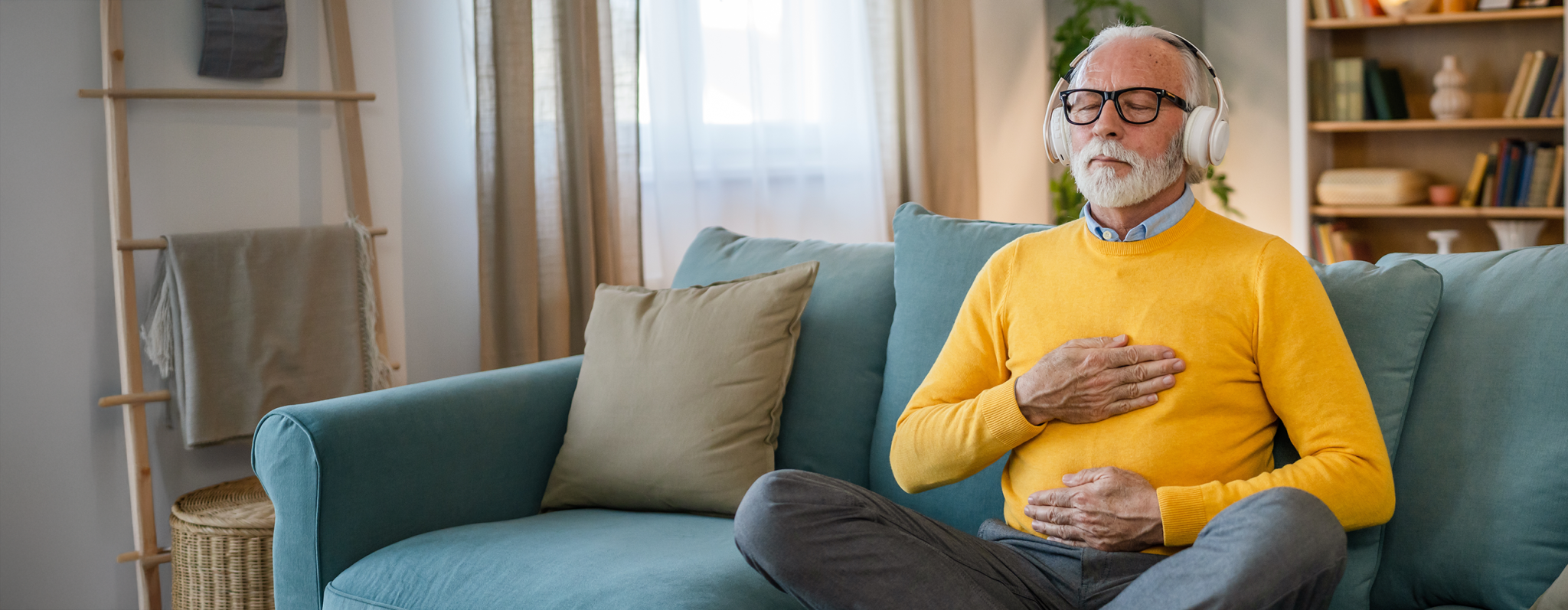 Ein älterer Mann mit weißen Haaren und Bart sitzt entspannt auf einem Sofa, trägt Kopfhörer und ein gelbes Sweatshirt. Er hat die Augen geschlossen und legt eine Hand auf die Brust und eine auf den Bauch, während er tief atmet oder meditiert.