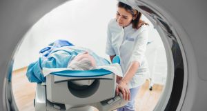 Blick aus einem Computertomographen: Eine Patientin oder ein Patient liegt für eine CT-Untersuchung auf dem Untersuchungstisch, eine medizinische Fachkraft steht daneben.