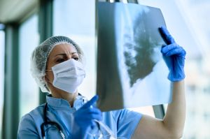 Eine Ärztin oder Pflegekraft mit Mundschutz, Haube und blauen Handschuhen betrachtet eine Röntgenaufnahme. Sie hält das Bild gegen das Licht, um Details besser erkennen zu können.