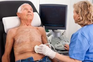 Eine medizinische Fachkraft führt bei einem älteren Mann eine Ultraschalluntersuchung im Brust- oder Bauchbereich durch. Der Patient liegt entspannt auf einer Untersuchungsliege, im Hintergrund ist ein Ultraschallgerät zu sehen.