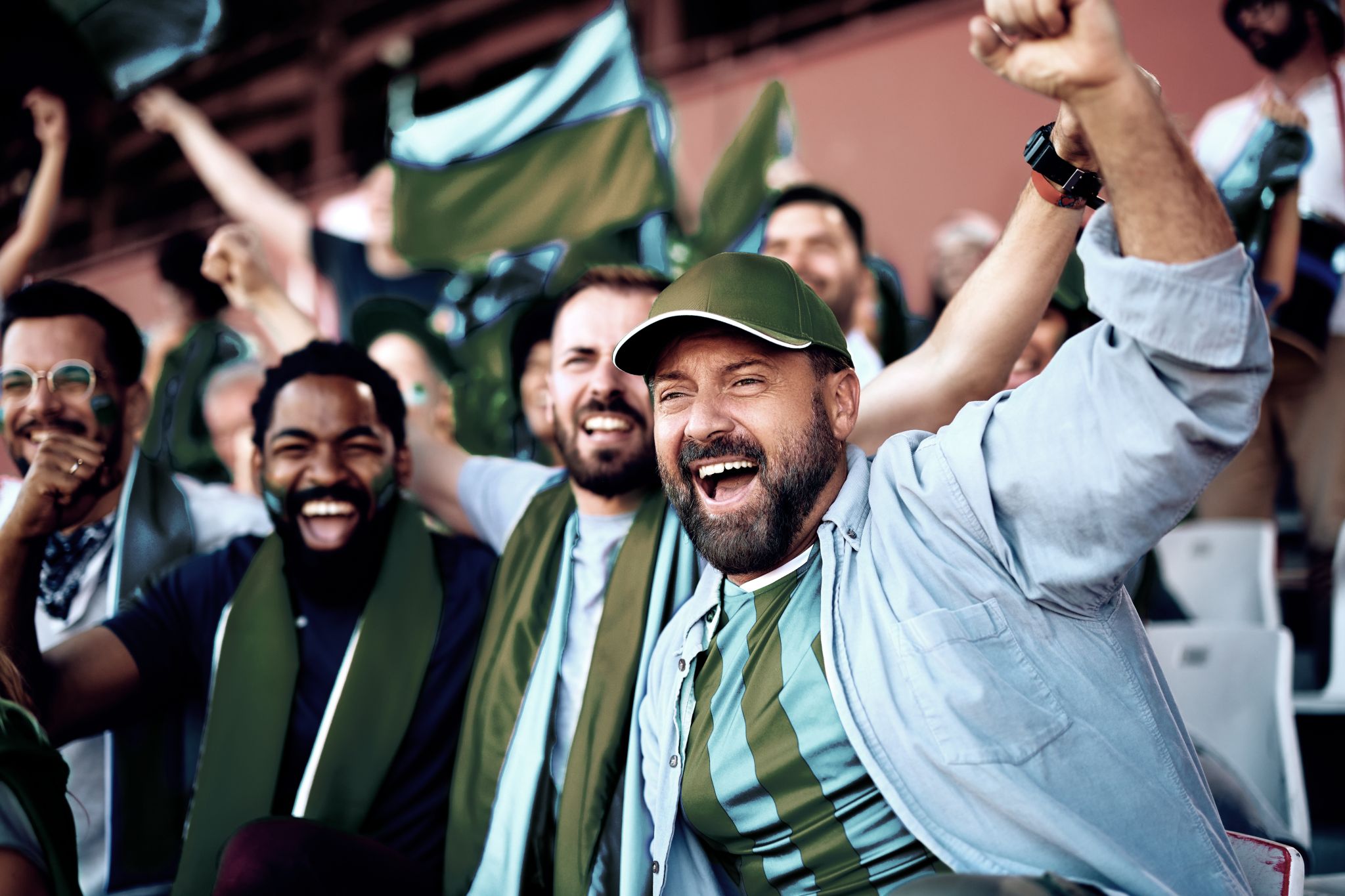 Eine Gruppe von Fußballfans im Stadion jubelt, hebt die Arme und schwenkt Fahnen in Grün- und Weißtönen.